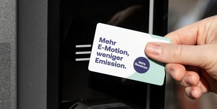 Hand hält eine Ladekarte mit der Aufschrift „Mehr E-Motion, weniger Emission.“ vor ein Kartenlesesymbol an der AC-Ladesäule, Schritt 1 der Ladeanleitung für AC-Laden.