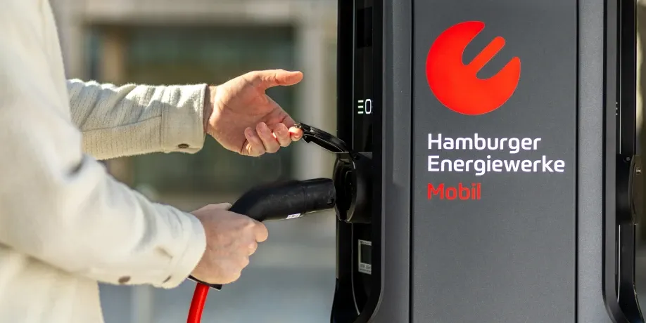 Eine Person steckt den Ladestecker mit rotem Kabel in die Buchse einer AC-Ladesäule mit dem Logo „Hamburger Energiewerke Mobil“, Schritt 2 der Ladeanleitung für AC-Laden.