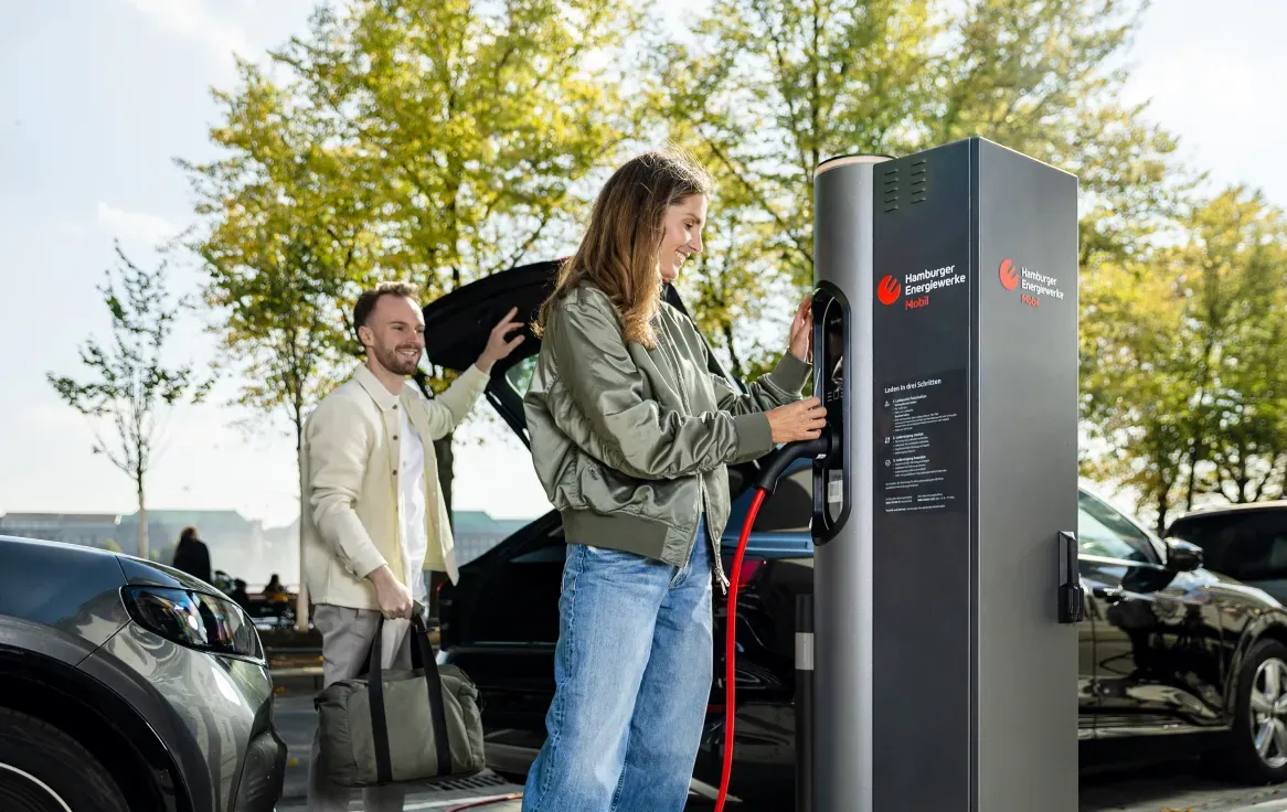 Eine Frau schließt ein Elektroauto an einer öffentlichen Ladesäule der Hamburger Energiewerke Mobil an, während ein Mann lächelnd mit Tasche daneben steht; im Hintergrund Bäume und parkende Autos an einem sonnigen Tag.