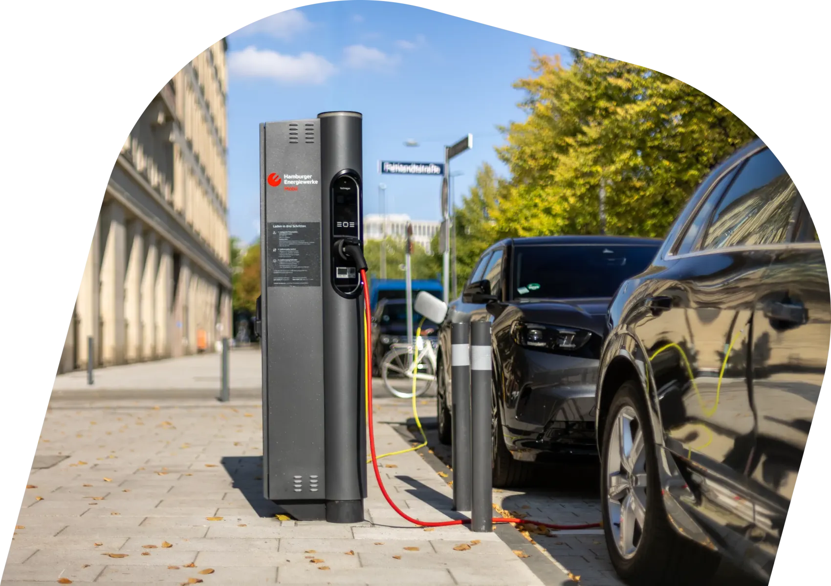 Öffentliche Ladesäule an einer Hamburger Straßenkante, ein Elektroauto lädt über ein rotes Kabel, im Hintergrund Bäume und Gebäude bei Sonnenschein; daneben der Text „Öffentliches Laden“.