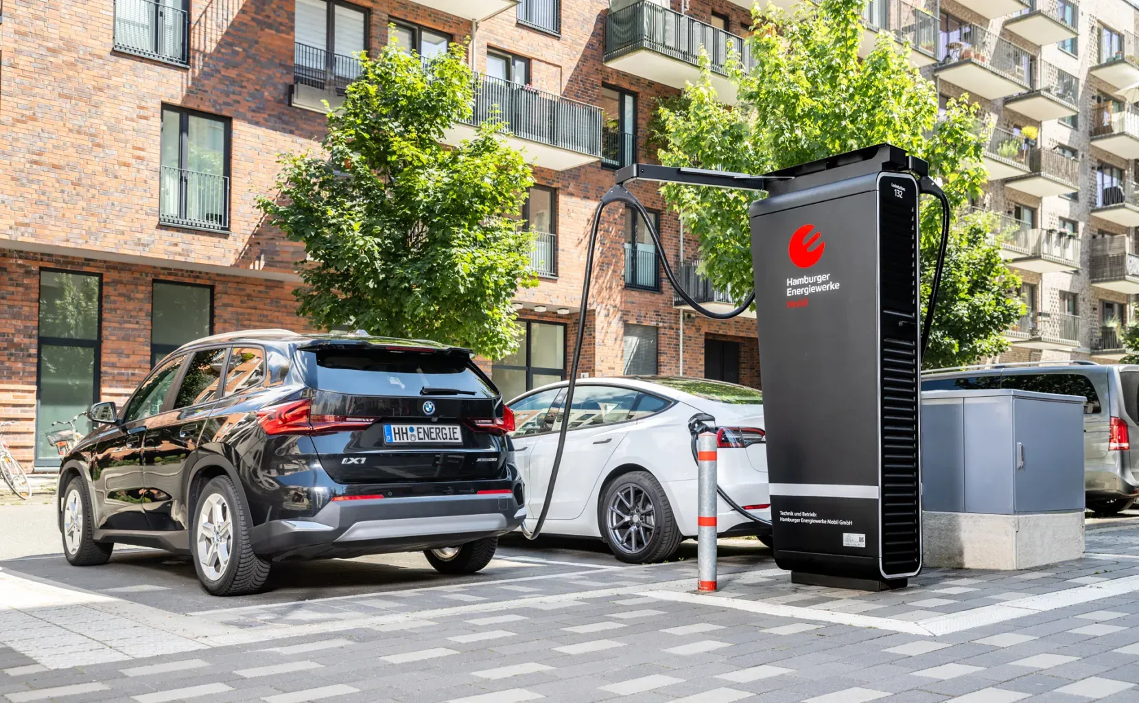 Zwei Elektroautos laden an einer großen schwarzen Schnellladesäule mit Hamburger Energiewerke Mobil-Logo in einer modernen Wohnstraße mit Bäumen und Mehrfamilienhäusern.