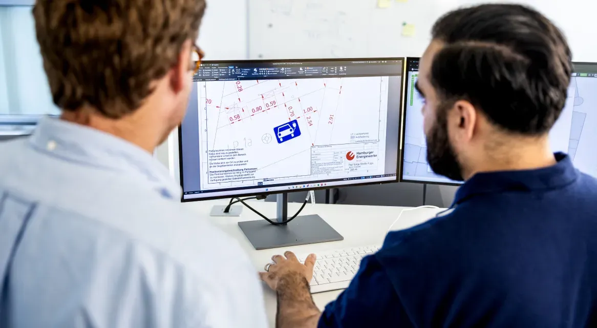 Zwei Mitarbeitende betrachten am Schreibtisch einen Bildschirm mit einem technischen Stadtplan und Planungsskizzen für Ladeinfrastruktur.