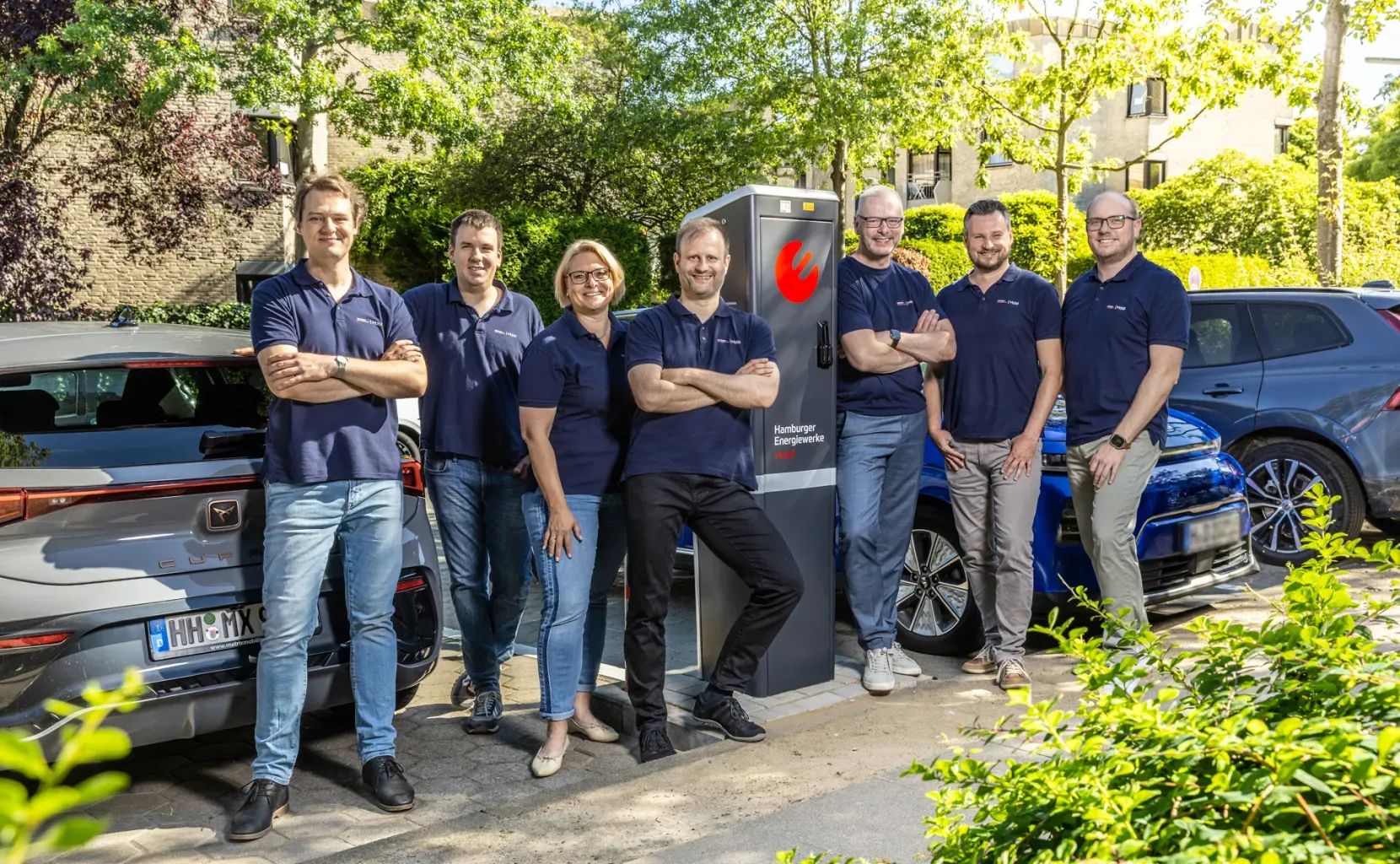 Mehrere Mitarbeitende in blauen Poloshirts stehen lächelnd um eine öffentliche Ladesäule der Hamburger Energiewerke Mobil und Elektroautos im Hintergrund in einer grünen Wohnstraße.
