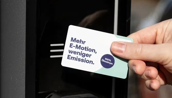 Hand hält eine Ladekarte mit der Aufschrift „Mehr E-Motion, weniger Emission.“ vor ein Kartenlesesymbol an der AC-Ladesäule, Schritt 1 der Ladeanleitung für AC-Laden.