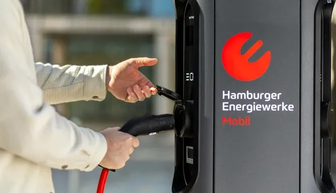 Eine Person steckt den Ladestecker mit rotem Kabel in die Buchse einer AC-Ladesäule mit dem Logo „Hamburger Energiewerke Mobil“, Schritt 2 der Ladeanleitung für AC-Laden.
