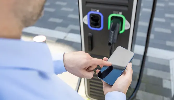 Person steht an einer DC-Ladesäule und bedient mit der rechten Hand ein Smartphone, Schritt 1 der Ladeanleitung für DC-Laden.