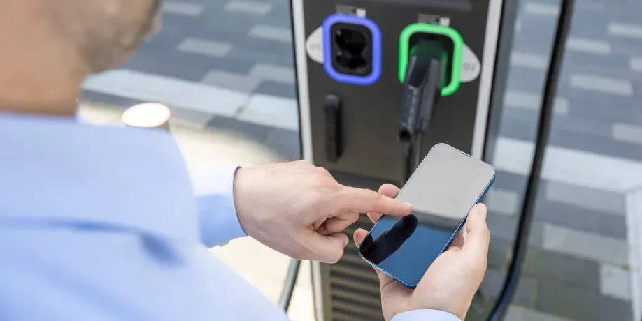 Person steht an einer DC-Ladesäule und bedient mit der rechten Hand ein Smartphone, Schritt 1 der Ladeanleitung für DC-Laden.