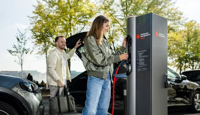 Eine Frau schließt ein Elektroauto an einer öffentlichen Ladesäule der Hamburger Energiewerke Mobil an, während ein Mann lächelnd mit Tasche daneben steht; im Hintergrund Bäume und parkende Autos an einem sonnigen Tag.
