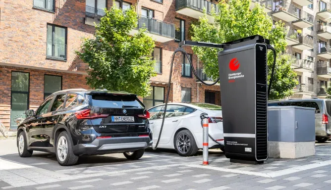 Zwei Elektroautos laden an einer großen schwarzen Schnellladesäule mit Hamburger Energiewerke Mobil-Logo in einer modernen Wohnstraße mit Bäumen und Mehrfamilienhäusern.