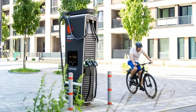 Moderne Schnellladesäule von Hamburger Energiewerke Mobil an einem breiten Stadtfußweg vor neuen Wohngebäuden, daneben fährt ein Radfahrer vorbei.
