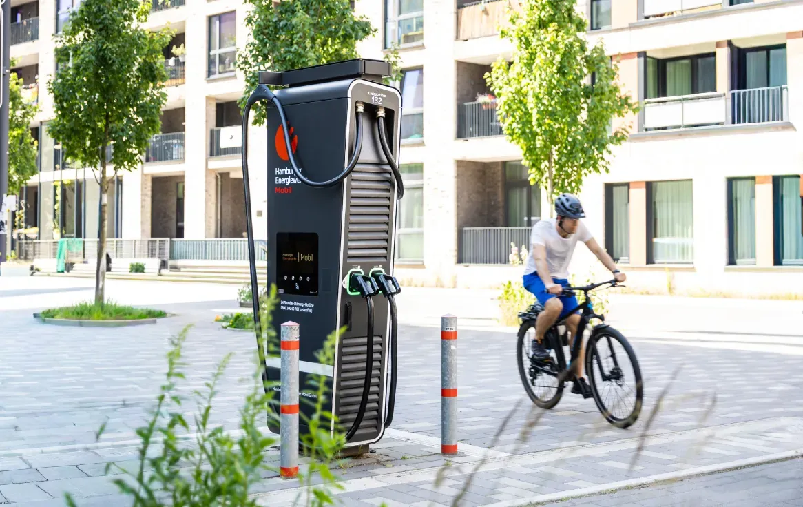 Moderne Schnellladesäule von Hamburger Energiewerke Mobil an einem breiten Stadtfußweg vor neuen Wohngebäuden, daneben fährt ein Radfahrer vorbei.