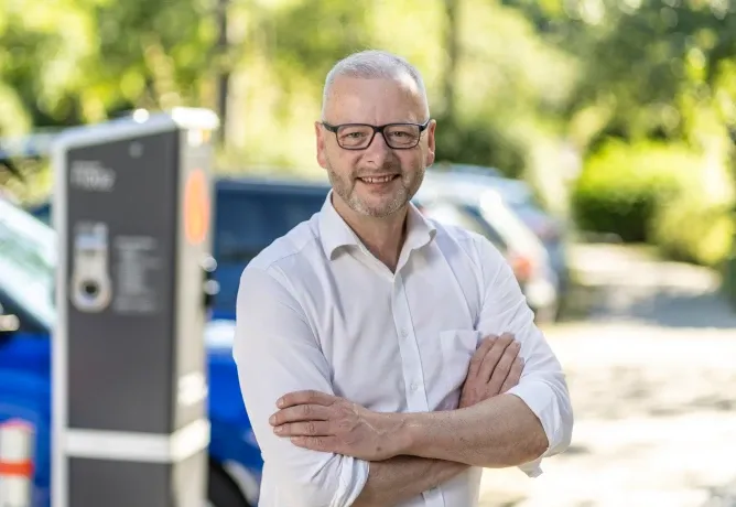 Mitarbeiter in weißem Hemd steht mit verschränkten Armen im Freien, im Hintergrund eine Ladesäule, parkende Fahrzeuge und Bäume.