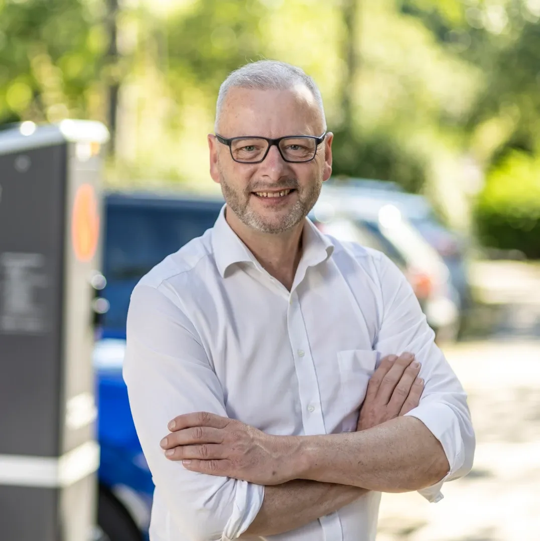 Mitarbeiter in weißem Hemd steht mit verschränkten Armen im Freien, im Hintergrund eine Ladesäule, parkende Fahrzeuge und Bäume.