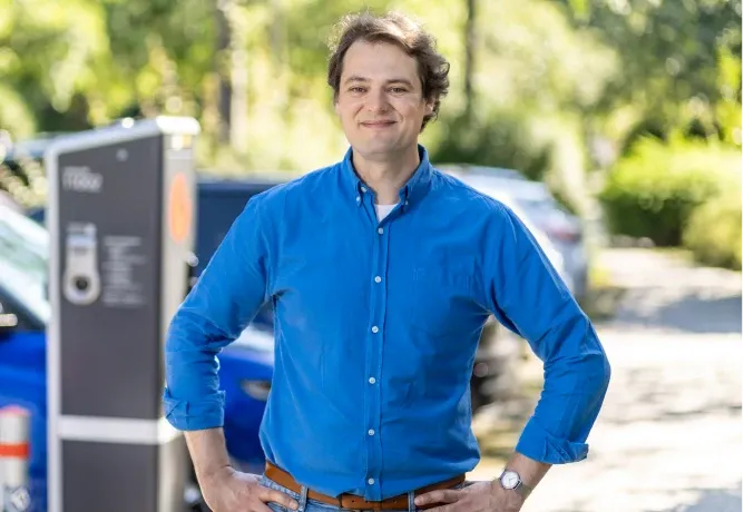 Mitarbeiter in blauem Hemd steht mit den Händen in die Hüften gestützt im Freien, im Hintergrund sind eine Ladesäule, parkende Fahrzeuge und Bäume zu sehen.