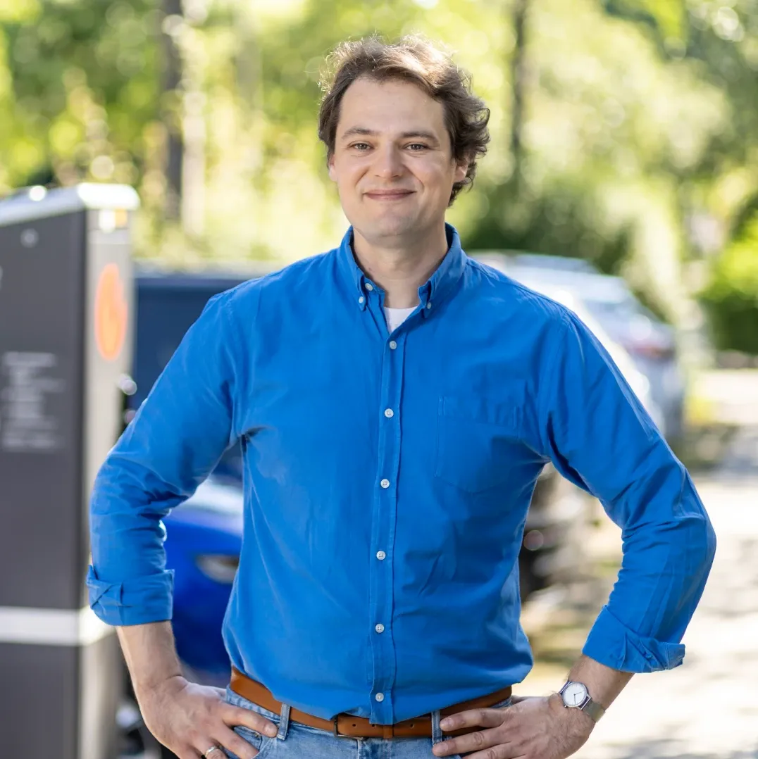 Mitarbeiter in blauem Hemd steht mit den Händen in die Hüften gestützt im Freien, im Hintergrund sind eine Ladesäule, parkende Fahrzeuge und Bäume zu sehen.