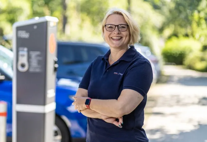 Mitarbeiterin in dunkelblauem Poloshirt mit verschränkten Armen steht im Freien neben einer Ladesäule, im Hintergrund sind ein blaues Auto und Bäume zu sehen.