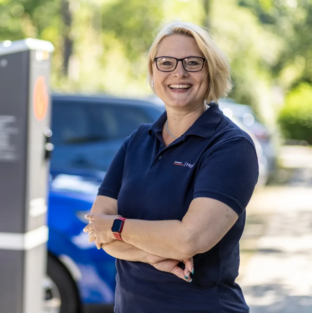 Mitarbeiterin in dunkelblauem Poloshirt mit verschränkten Armen steht im Freien neben einer Ladesäule, im Hintergrund sind ein blaues Auto und Bäume zu sehen.