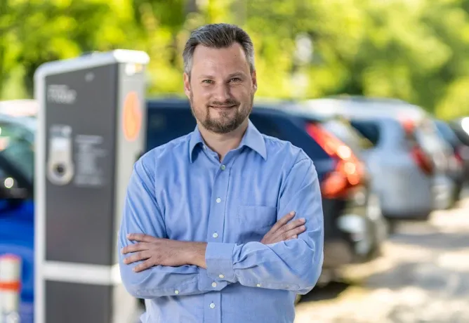 Mitarbeiter in hellblauem Hemd mit verschränkten Armen steht im Freien vor einer Ladesäule, im Hintergrund parken mehrere Fahrzeuge an einer begrünten Straße.
