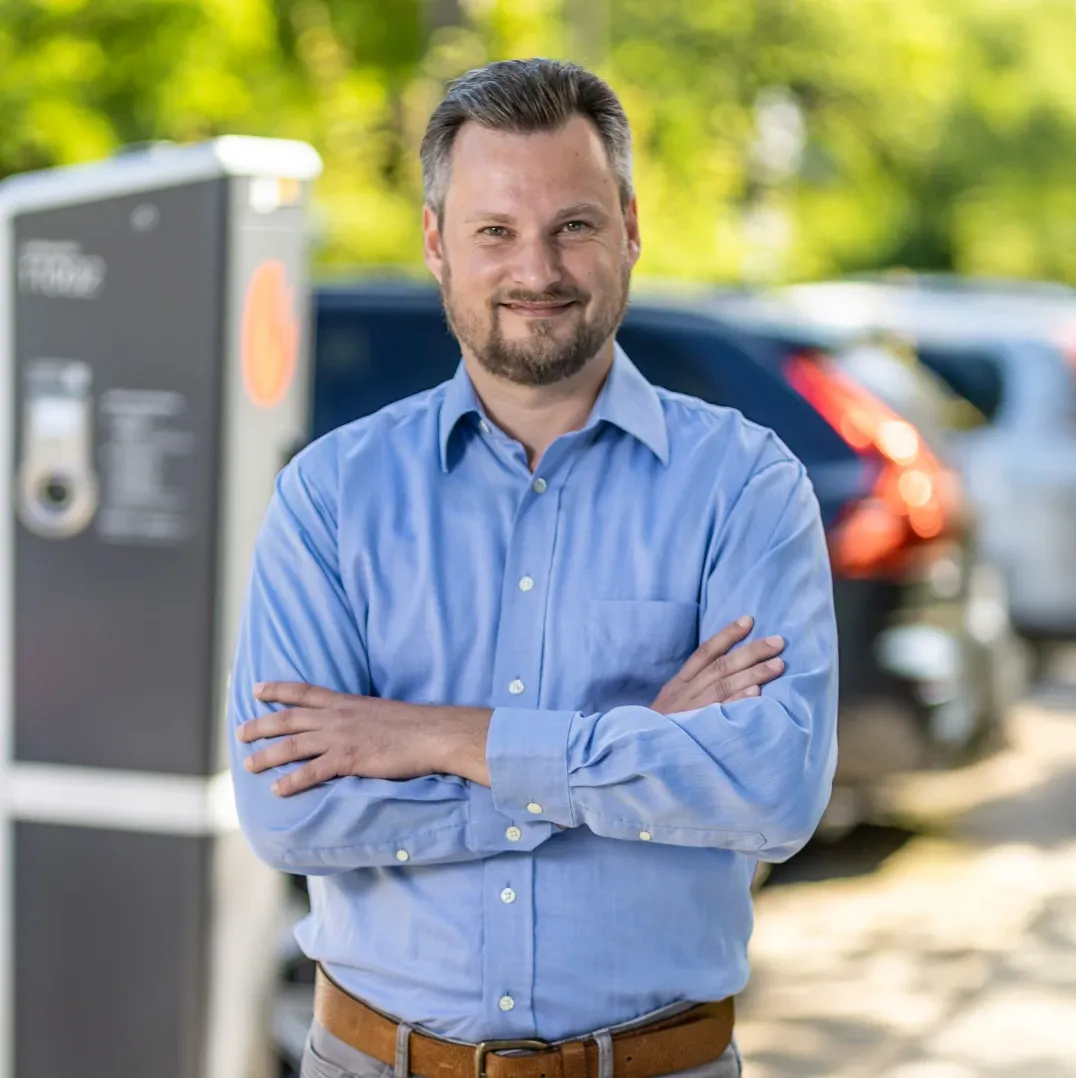 Mitarbeiter in hellblauem Hemd mit verschränkten Armen steht im Freien vor einer Ladesäule, im Hintergrund parken mehrere Fahrzeuge an einer begrünten Straße.