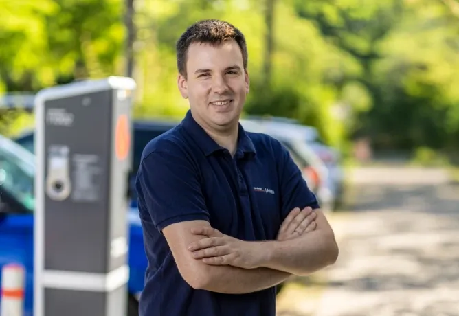 Mitarbeiter in dunklem Poloshirt mit verschränkten Armen steht im Freien neben einer öffentlichen Ladesäule, im Hintergrund sind Bäume und parkende Autos zu sehen.