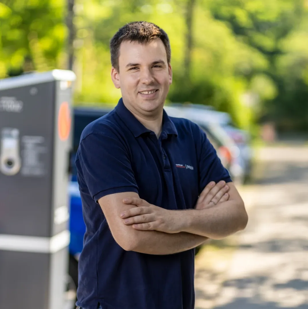 Mitarbeiter in dunklem Poloshirt mit verschränkten Armen steht im Freien neben einer öffentlichen Ladesäule, im Hintergrund sind Bäume und parkende Autos zu sehen.