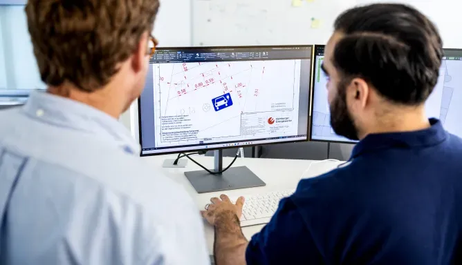 Zwei Mitarbeitende betrachten am Schreibtisch einen Bildschirm mit einem technischen Stadtplan und Planungsskizzen für Ladeinfrastruktur.