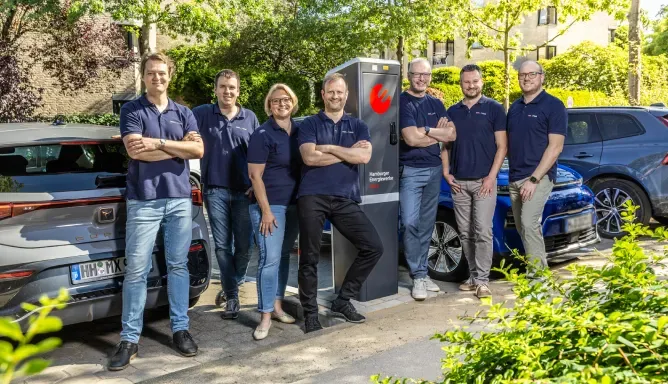 Mehrere Mitarbeitende in blauen Poloshirts stehen lächelnd um eine öffentliche Ladesäule der Hamburger Energiewerke Mobil und Elektroautos im Hintergrund in einer grünen Wohnstraße.
