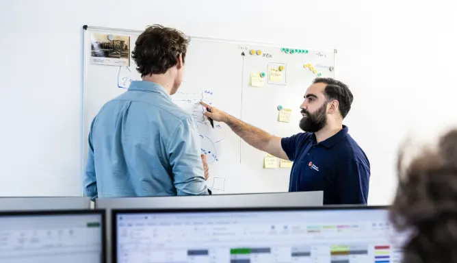 Zwei Mitarbeitende stehen vor einem Whiteboard mit Skizzen und Notizen und diskutieren technische Abläufe, im Vordergrund sind Computerbildschirme mit Tabellen und Diagrammen zu sehen.