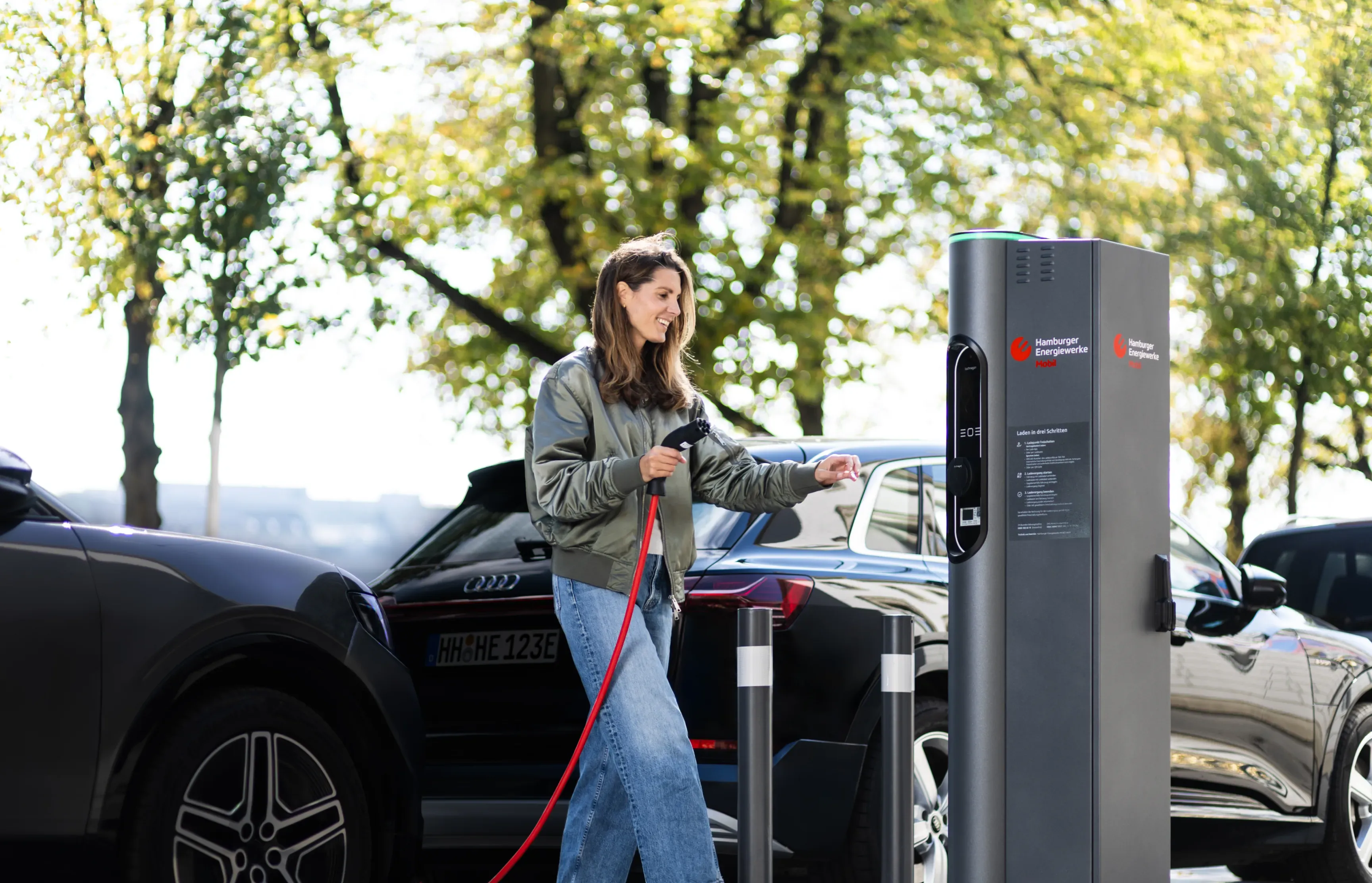 Person lädt ein Elektroauto an einer öffentlichen Ladesäule von Hamburg Energie, im Hintergrund stehen weitere Fahrzeuge. Text im Bild: „Elektromobilität für Hamburg“.