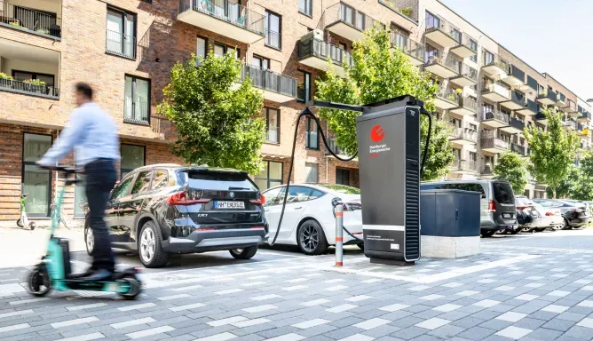 Elektroautos stehen an einem modernen Schnelllader von Hamburger Energiewerke Mobil in einer Wohnstraße, während im Vordergrund eine Person auf einem E-Scooter vorbeifährt.