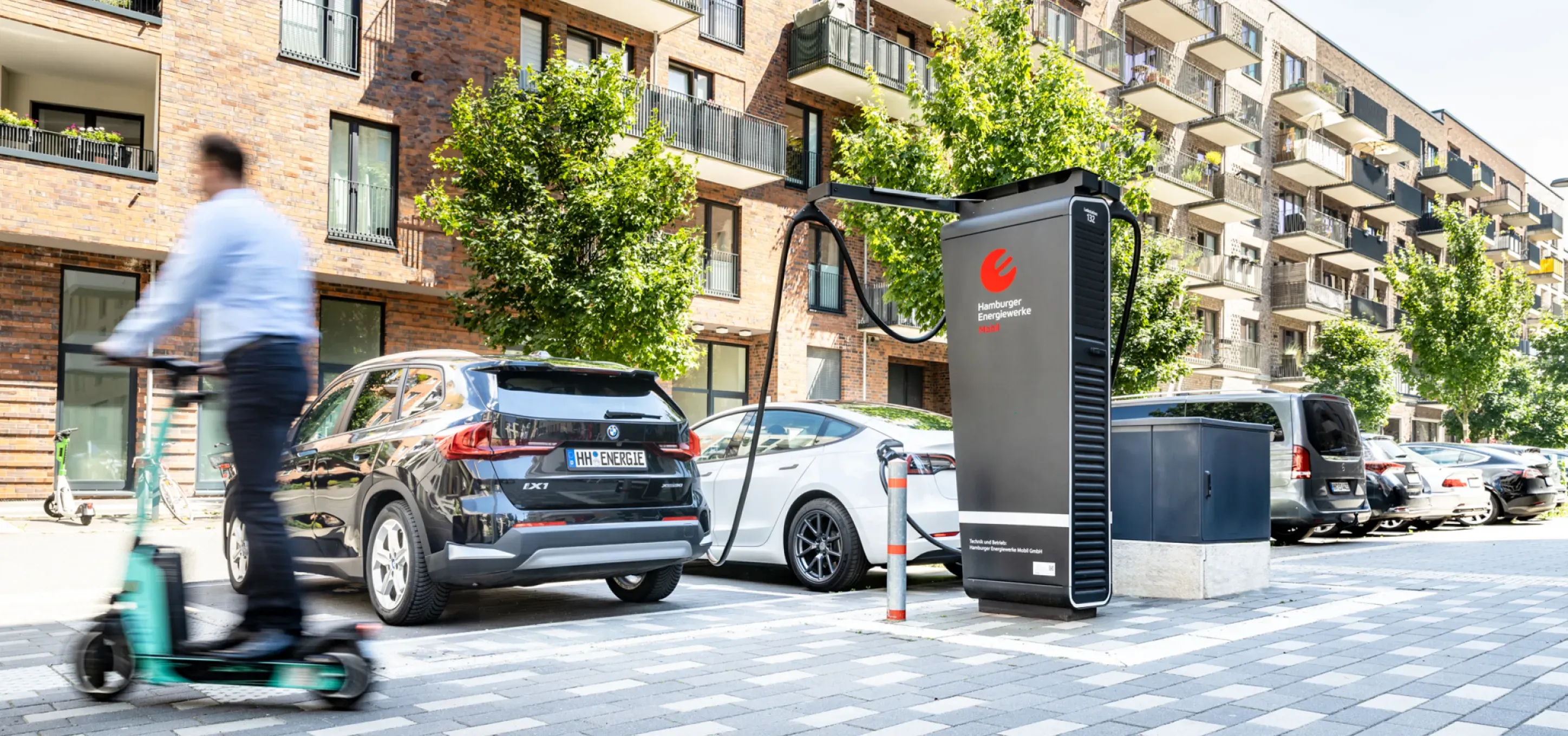 Elektroautos stehen an einem modernen Schnelllader von Hamburger Energiewerke Mobil in einer Wohnstraße, während im Vordergrund eine Person auf einem E-Scooter vorbeifährt.