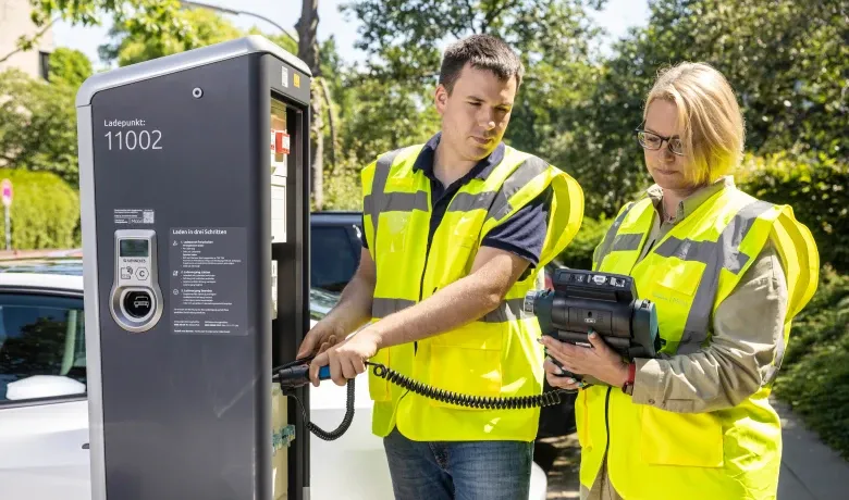Zwei Personen in gelben Warnwesten prüfen eine öffentliche Ladesäule; eine Person hält den Ladestecker, die andere ein mobiles Prüfgerät, im Hintergrund stehen Bäume und parkende Autos.