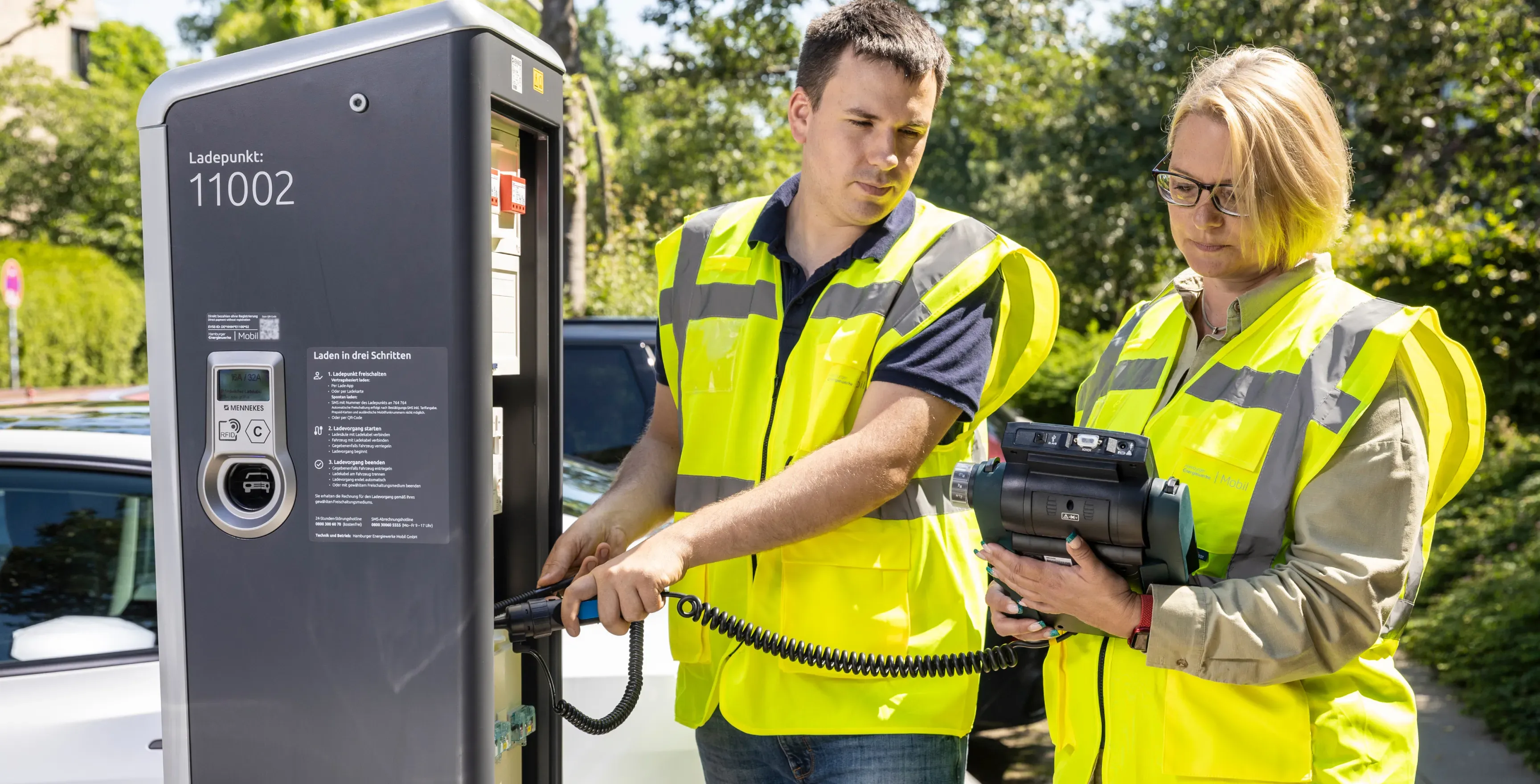 Zwei Personen in gelben Warnwesten prüfen eine öffentliche Ladesäule; eine Person hält den Ladestecker, die andere ein mobiles Prüfgerät, im Hintergrund stehen Bäume und parkende Autos.