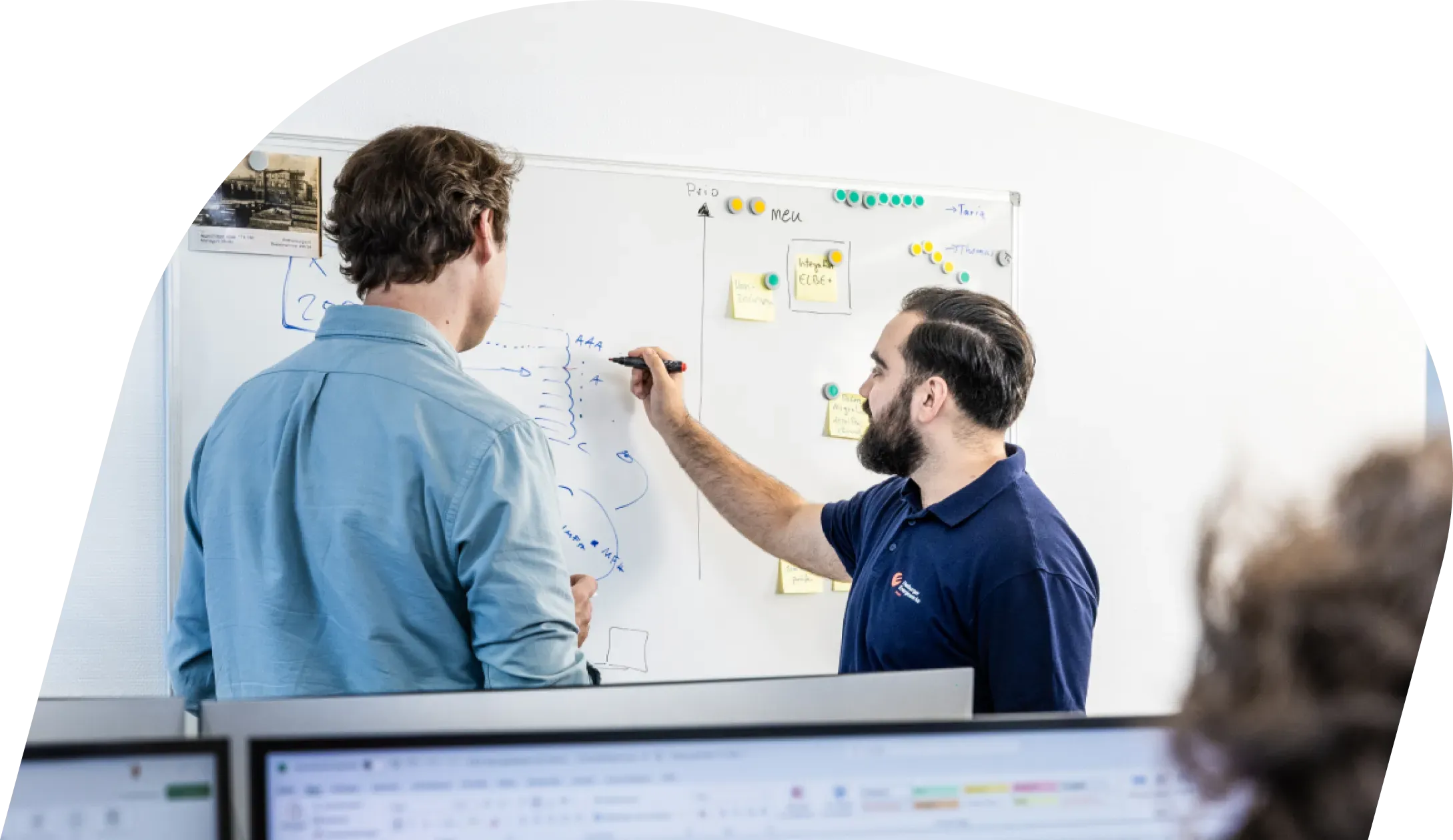 Zwei Mitarbeitende stehen vor einem Whiteboard mit Skizzen und Notizen und besprechen technische Abläufe, im Vordergrund sind Computerbildschirme zu sehen; daneben der Text „Du möchtest Teil unseres Teams werden?“ mit Button zur Karriereseite.