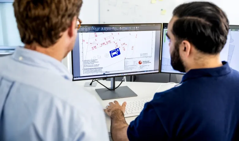 Zwei Mitarbeitende betrachten am Schreibtisch einen Bildschirm mit einem technischen Stadtplan und Planungsskizzen für Ladeinfrastruktur.