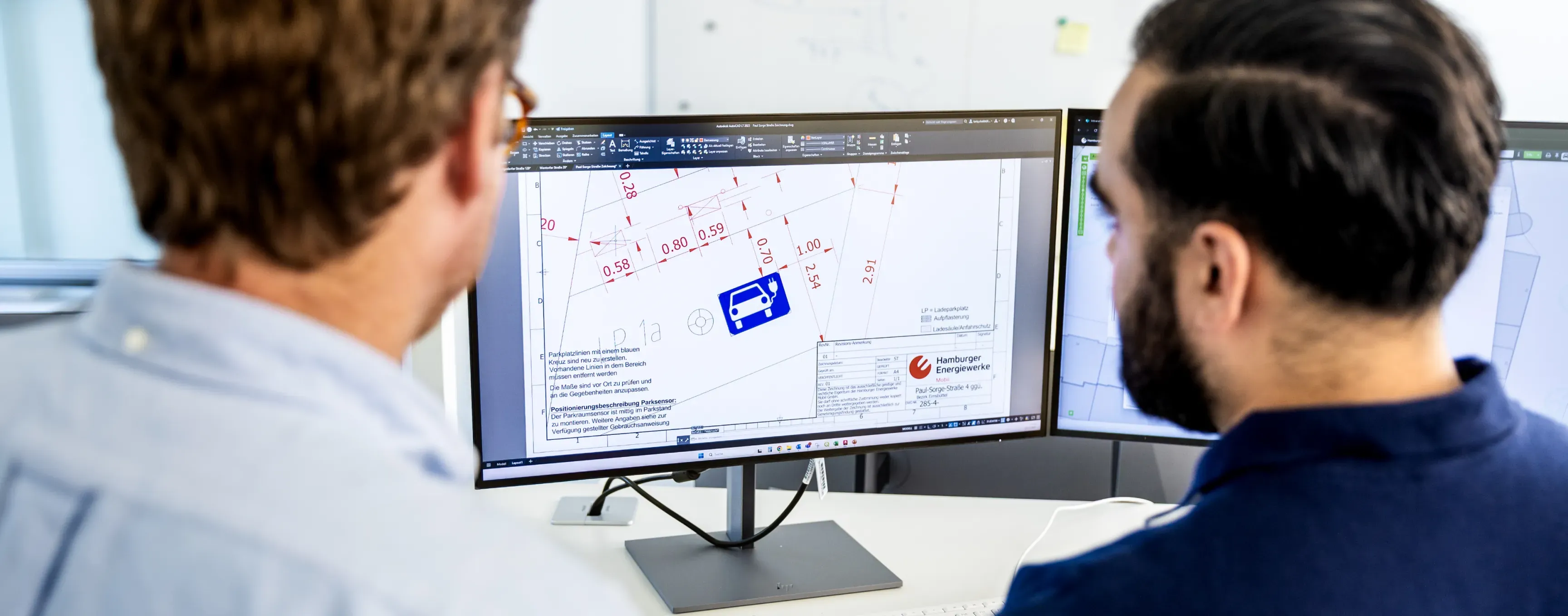 Zwei Mitarbeitende betrachten am Schreibtisch einen Bildschirm mit einem technischen Stadtplan und Planungsskizzen für Ladeinfrastruktur.
